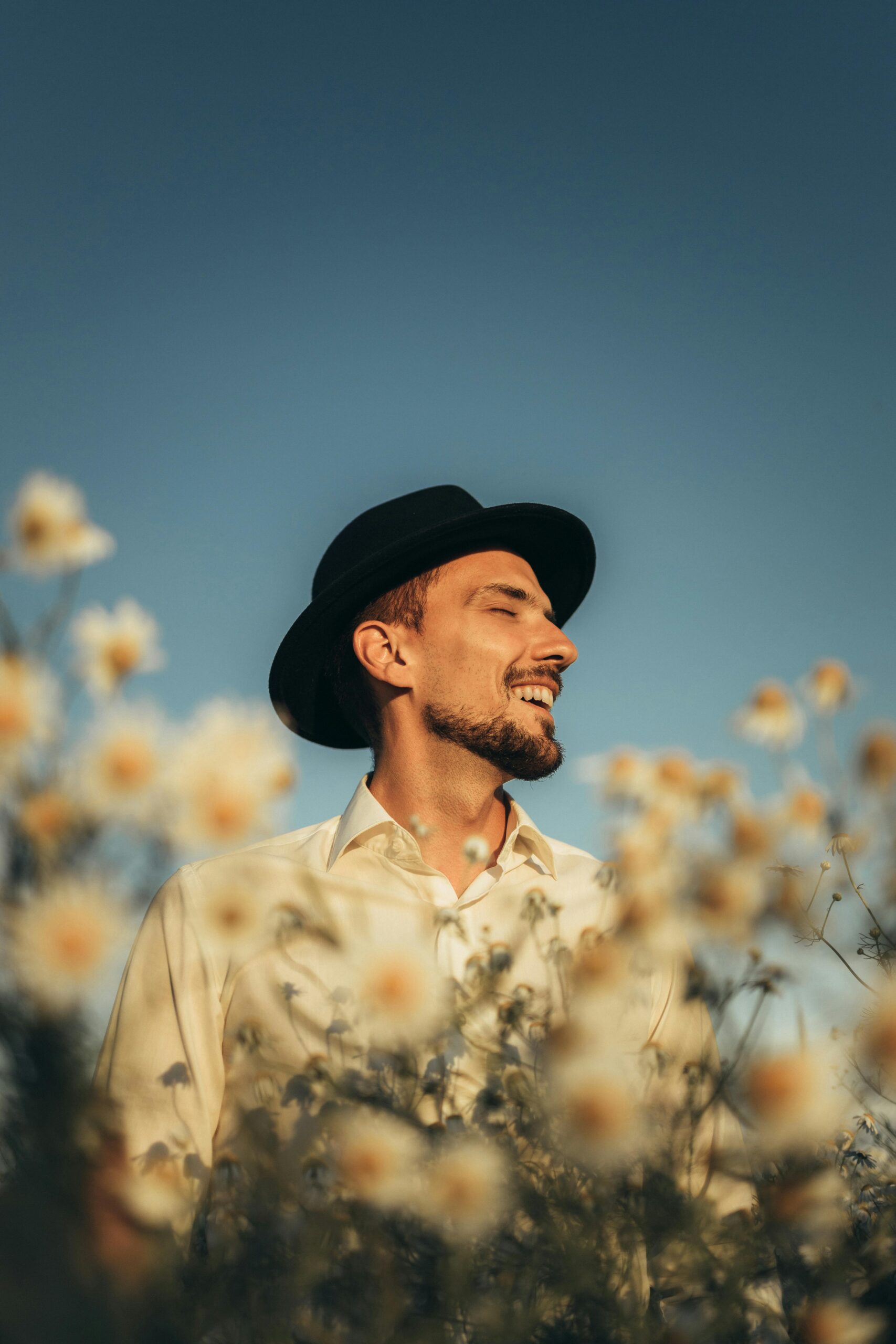 Smiling man with a fedora amidst flowers under a blue sky, exuding joy and tranquility.